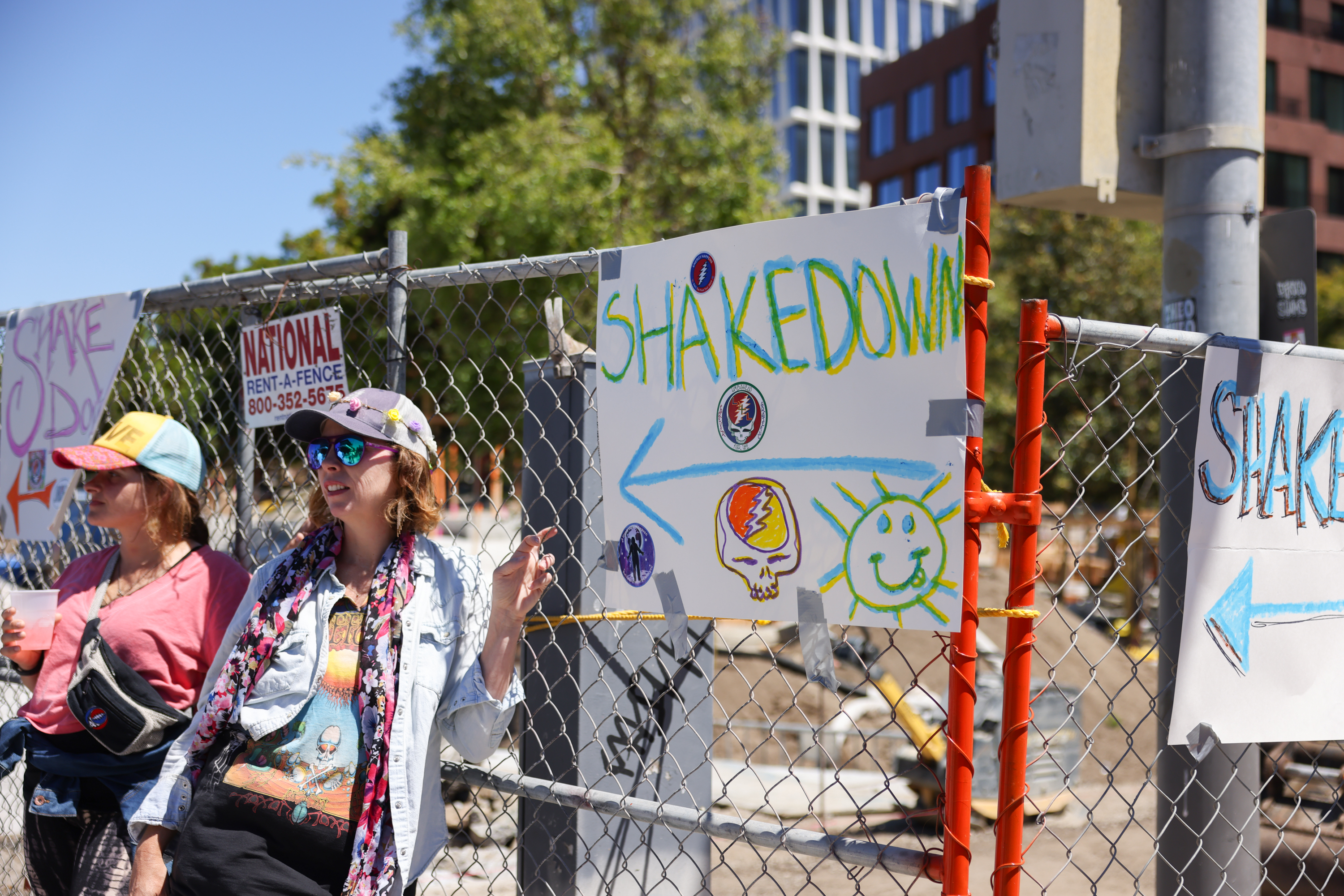 Two people stand by a chain-link fence covered with colorful "SHAKEDOWN" signs and doodles, including a skull and a smiling sun.
